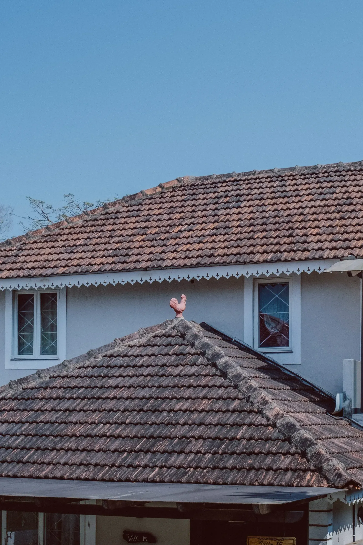 Traditional Indian house with terracotta tiled roof and decorative rooster weathervane under blue sky.