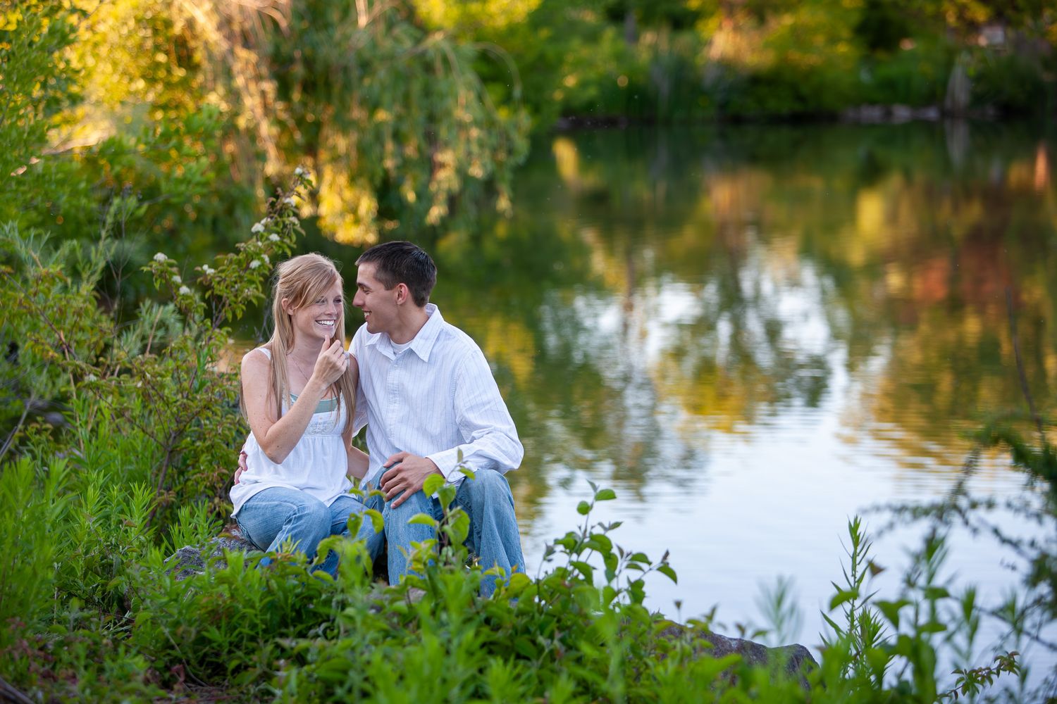 Romantic couple sitting together by a peaceful pond surrounded by lush green vegetation at sunset.