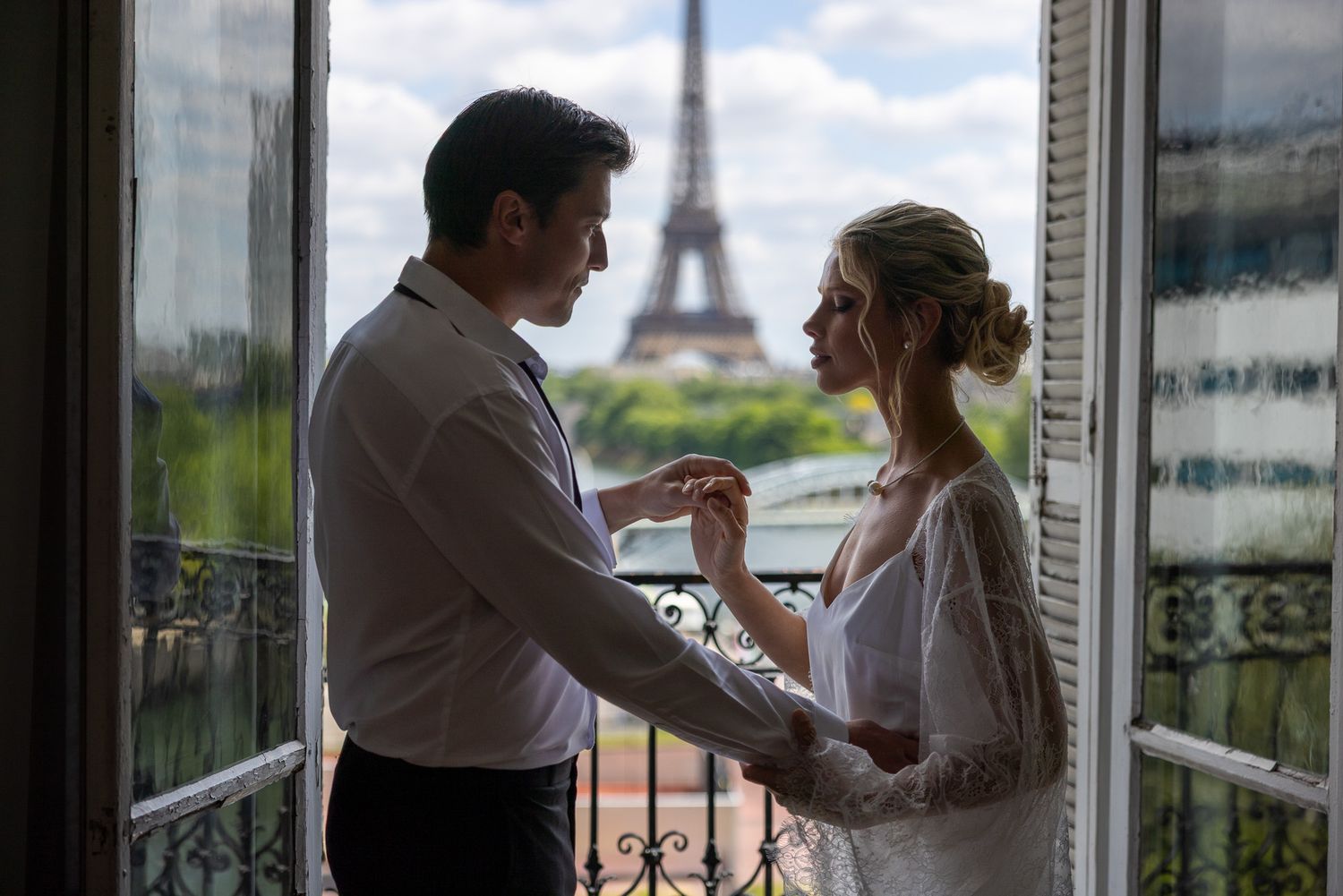 Couple holds hands on Paris balcony with Eiffel Tower visible between them.