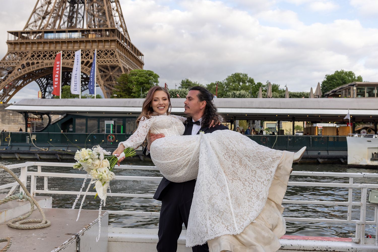 Groom carrying bride in wedding dress near the Eiffel Tower along the Seine river.