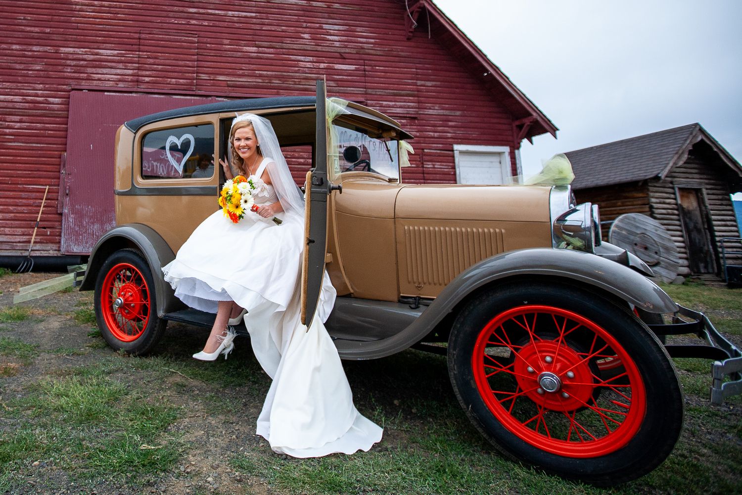 Bride in white gown holds yellow bouquet beside a vintage tan and red car near a red barn.