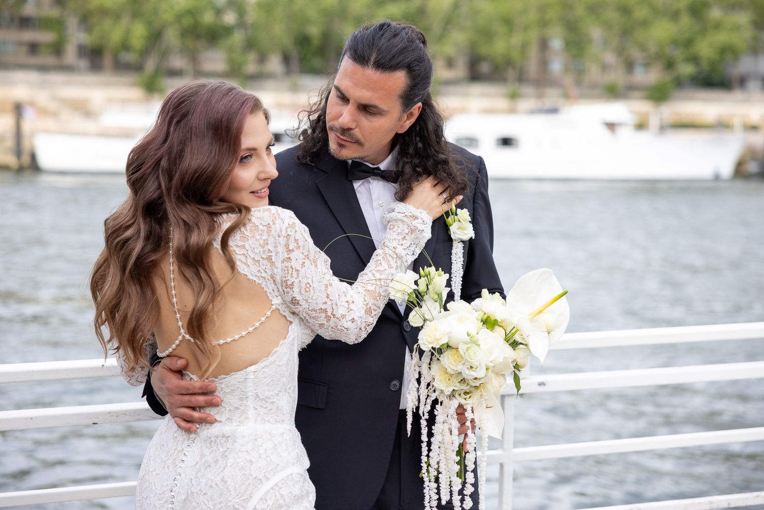 Bride and groom embrace on waterfront dock, bride holding white floral bouquet.