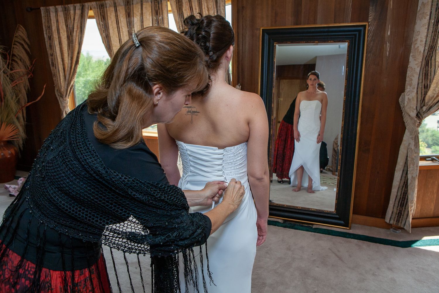 Woman helping bride fasten white wedding dress while reflected in large mirror.