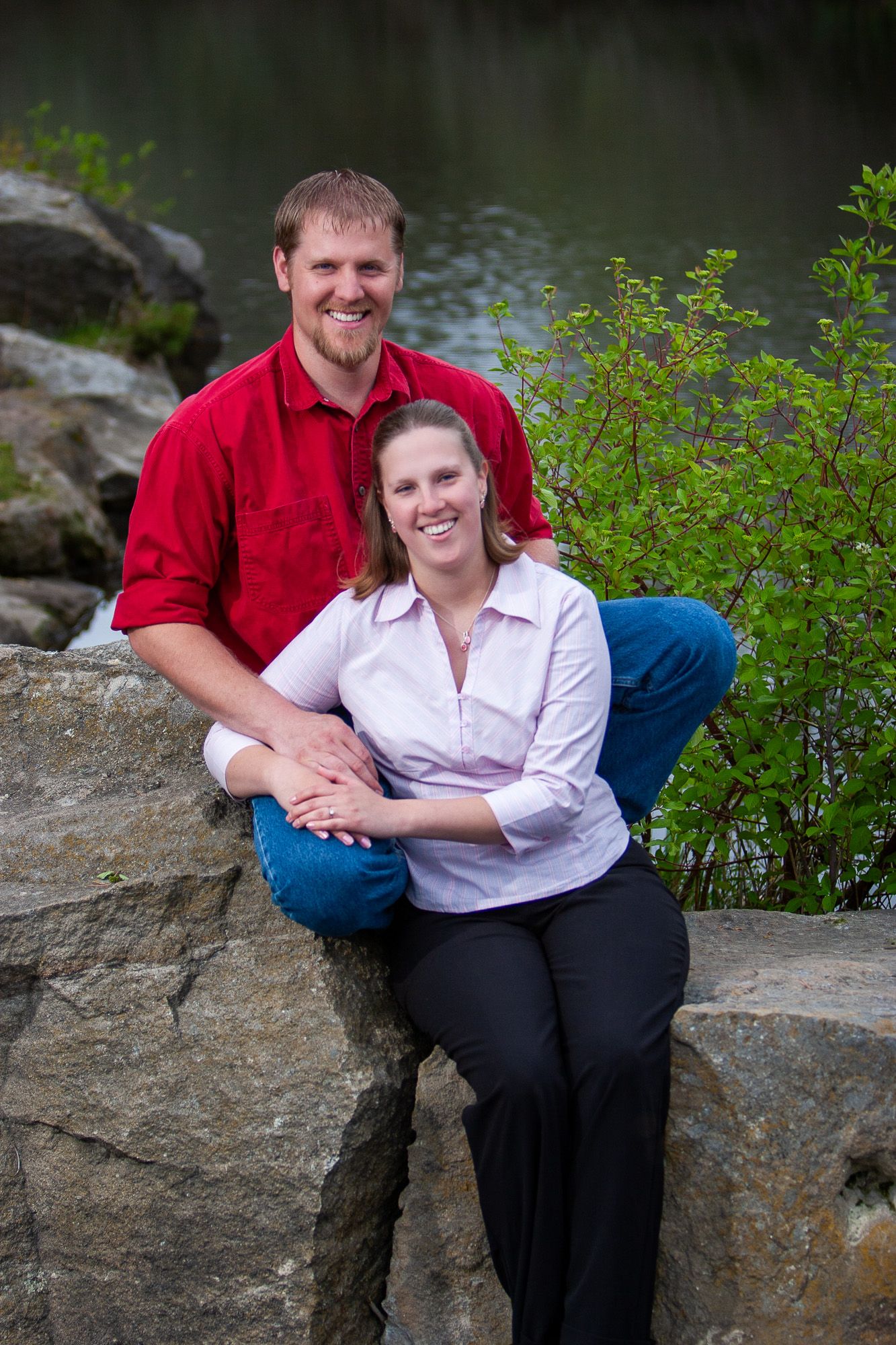 Happy couple posing on rocky terrain near a lake with lush green foliage behind them.