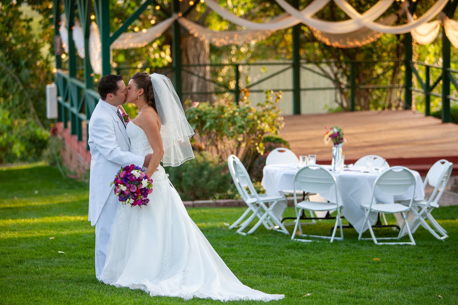 A bride and groom share a kiss on a lush green lawn with elegant white reception tables and a gazebo backdrop.