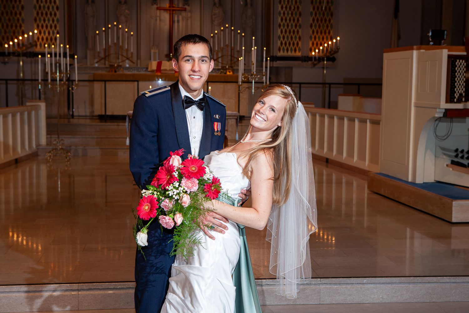 Groom in military uniform and bride in white gown with veil pose together inside a church.