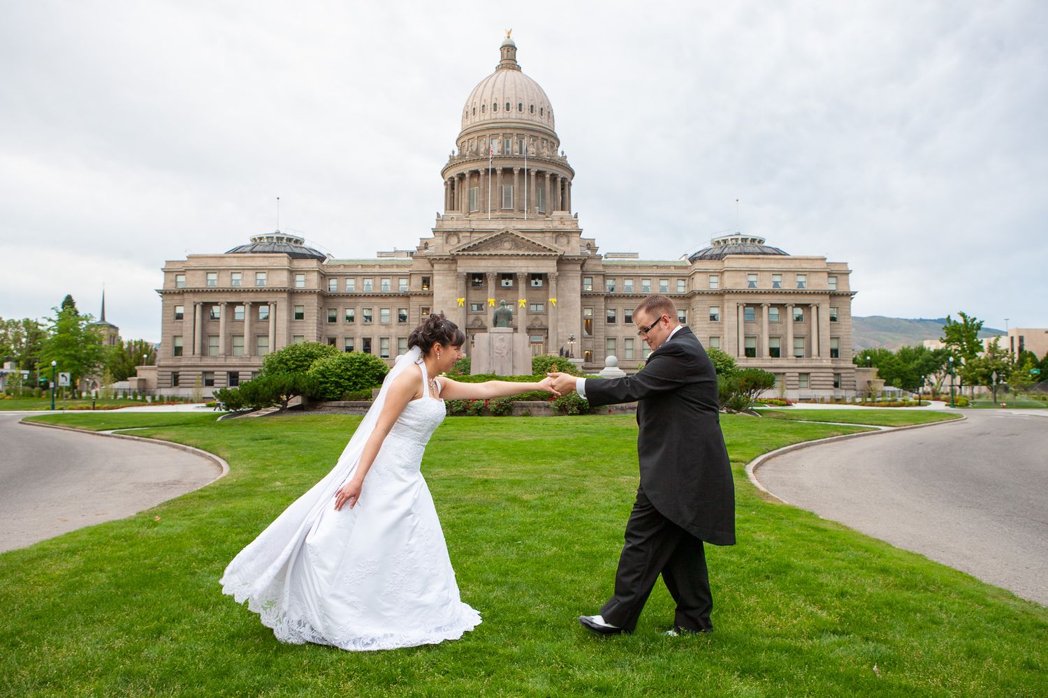 Bride and groom dance on green lawn in front of a grand state capitol building.