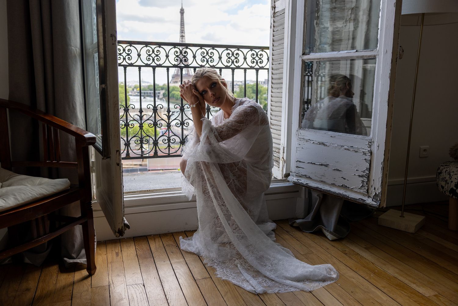 Bride in lace gown sits pensively on floor of Paris apartment balcony doorway.