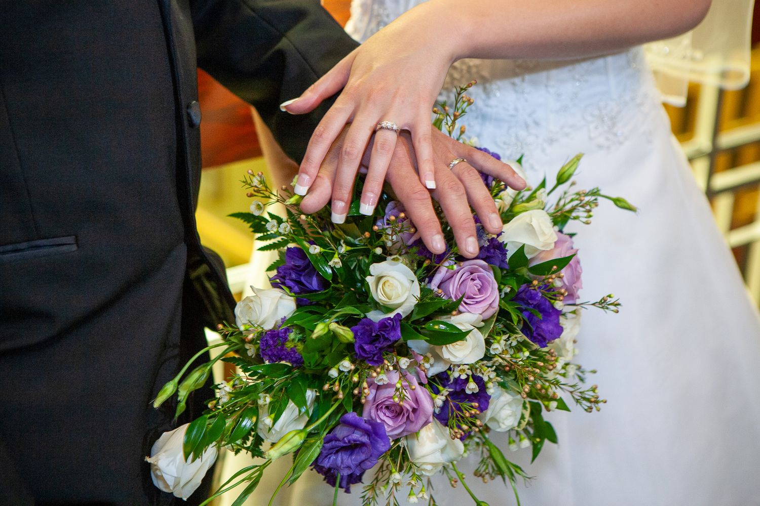 Bride and groom displaying wedding rings over a purple and white bridal bouquet.
