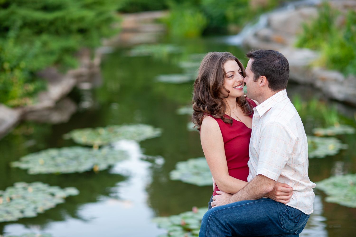 Romantic couple embracing and smiling near a tranquil lily pond during an outdoor engagement session.
