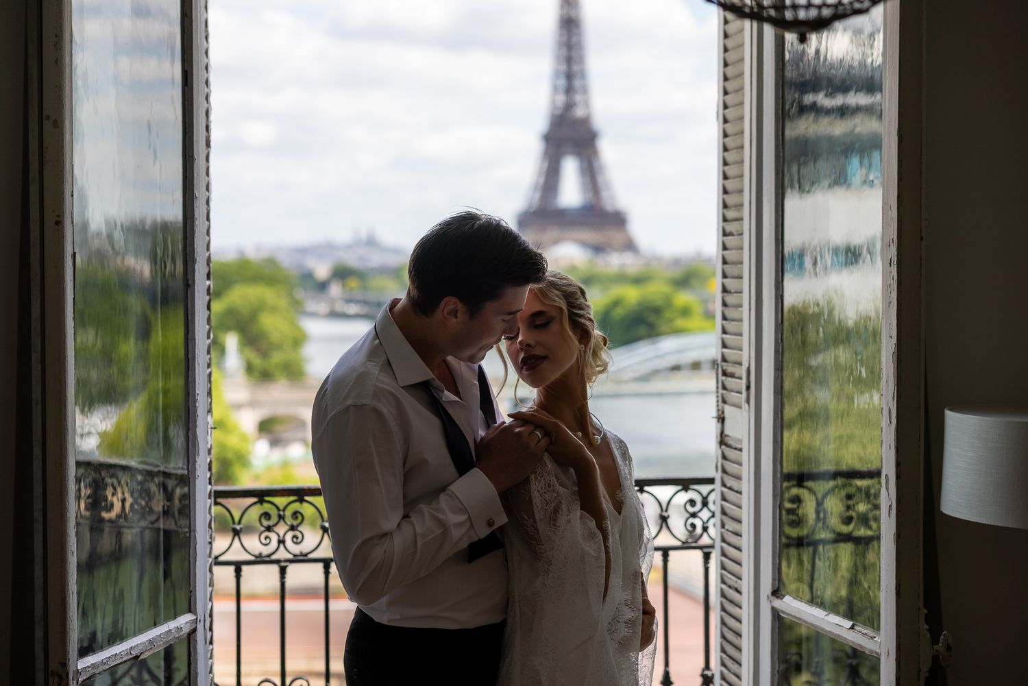 Newlyweds embrace intimately in silhouette framed by open balcony doors and Eiffel Tower.