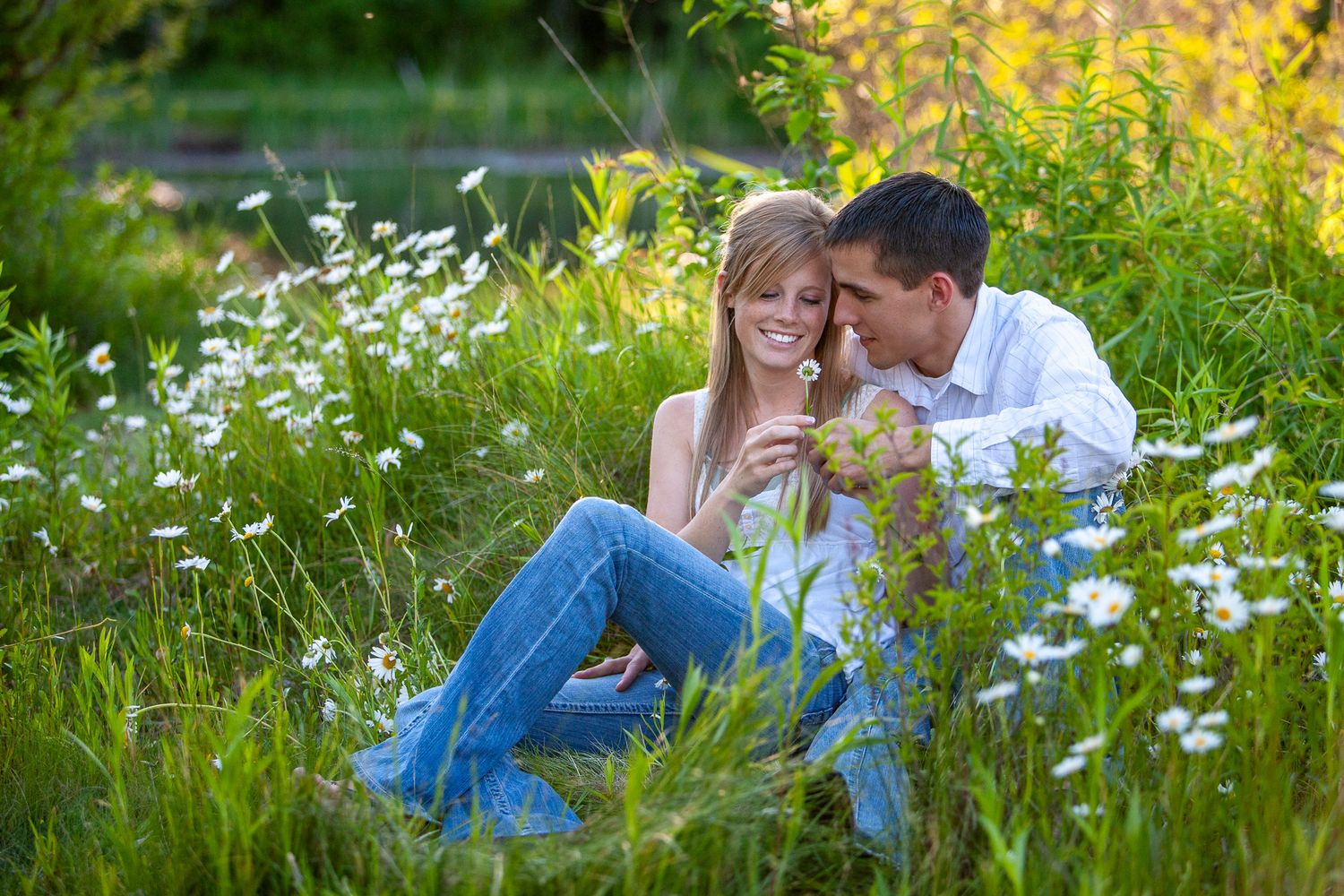 A romantic couple sitting together in a sunny wildflower meadow with daisies blooming around them.
