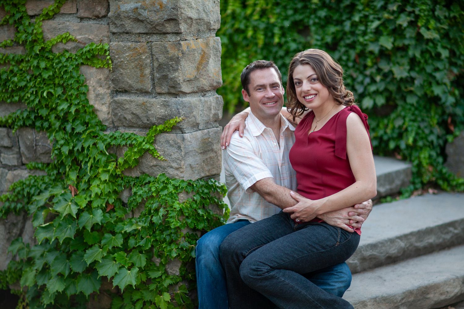 A smiling couple poses together against a stone wall covered in lush green ivy during an outdoor engagement photo session.