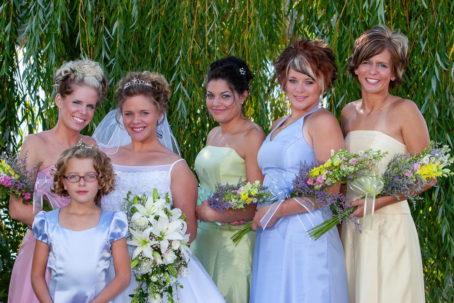 Bride with bridesmaids in pastel dresses holding floral bouquets outdoors under a willow tree.