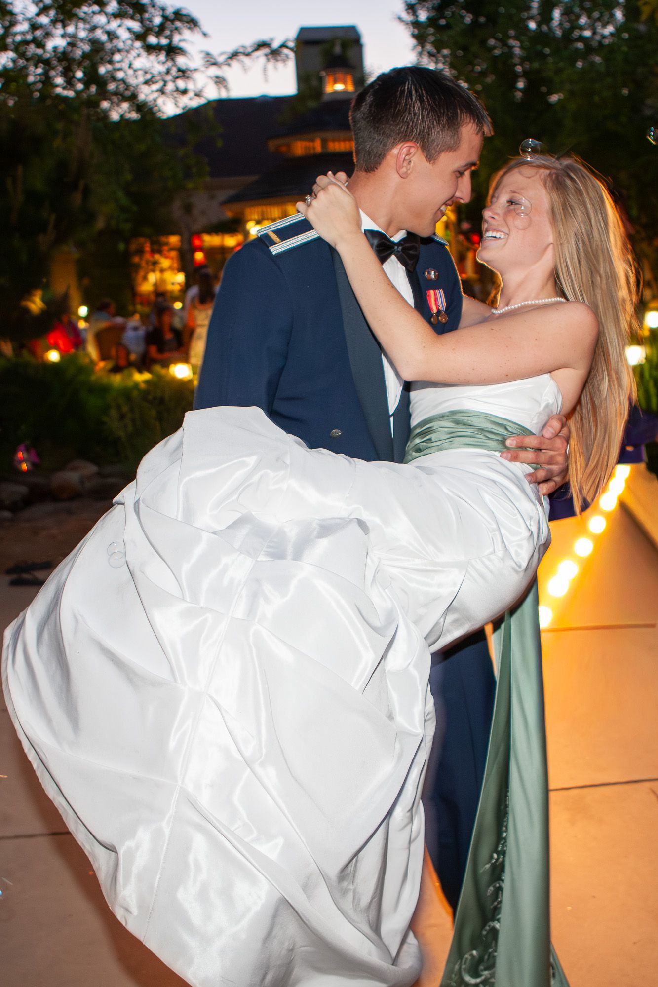 Military groom in uniform carrying his smiling bride in a white gown at an evening outdoor wedding.