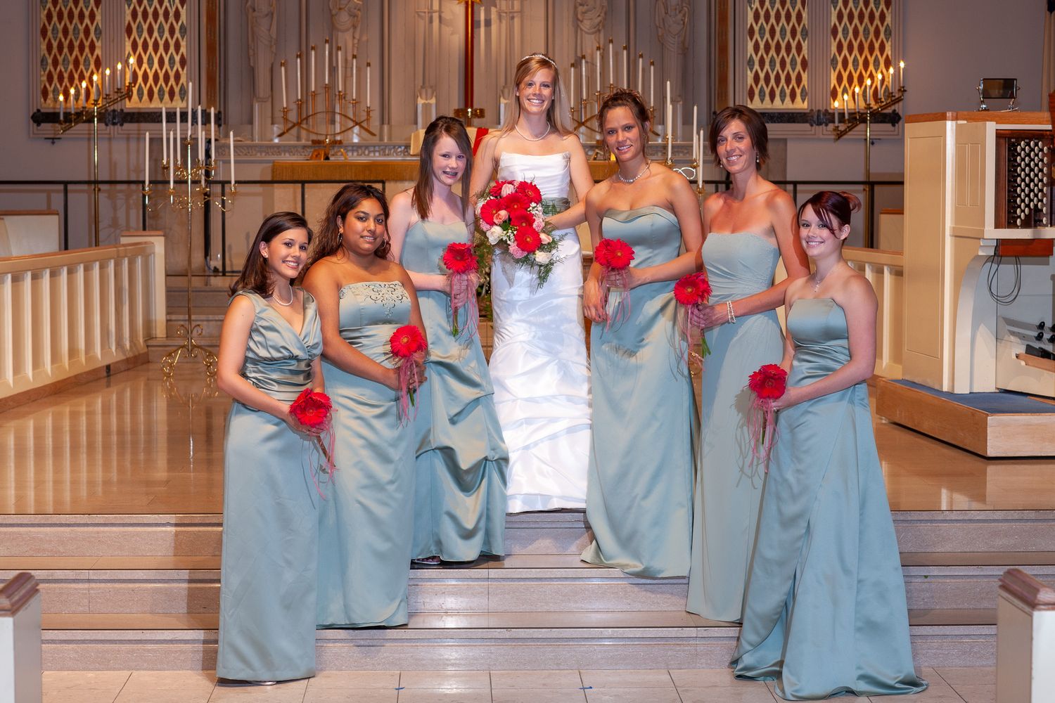 Bride in white gown poses with six bridesmaids in teal dresses holding red bouquets inside a church.