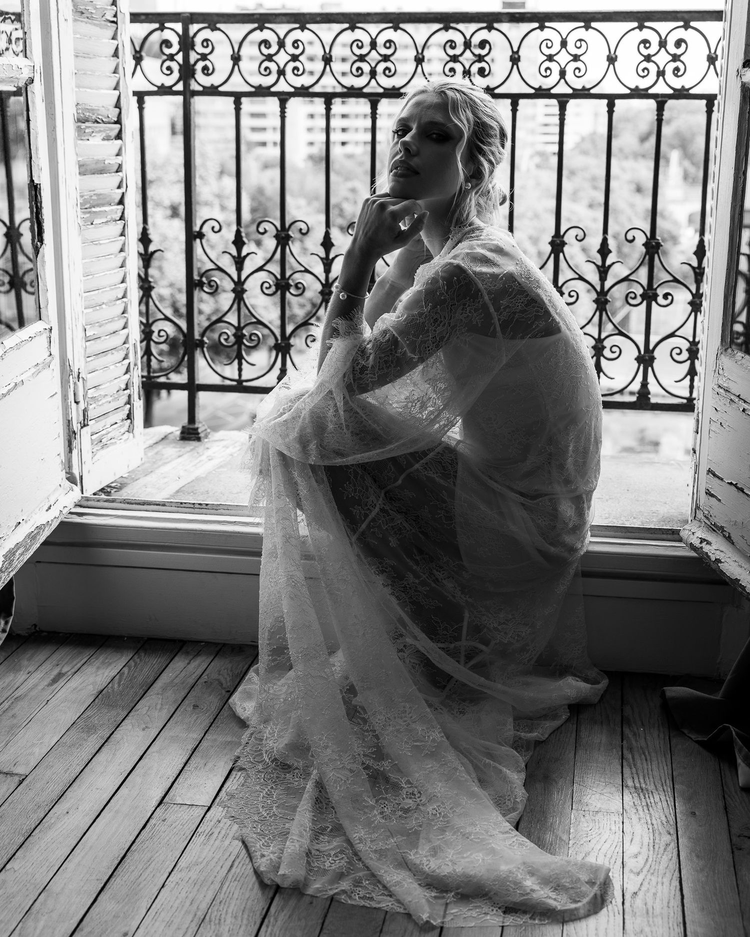 Bride in lace wedding gown sitting by ornate balcony window in black and white.
