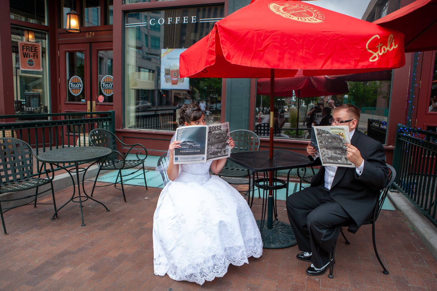 Bride and groom sit at outdoor coffee shop reading newspapers in wedding attire under a red umbrella.