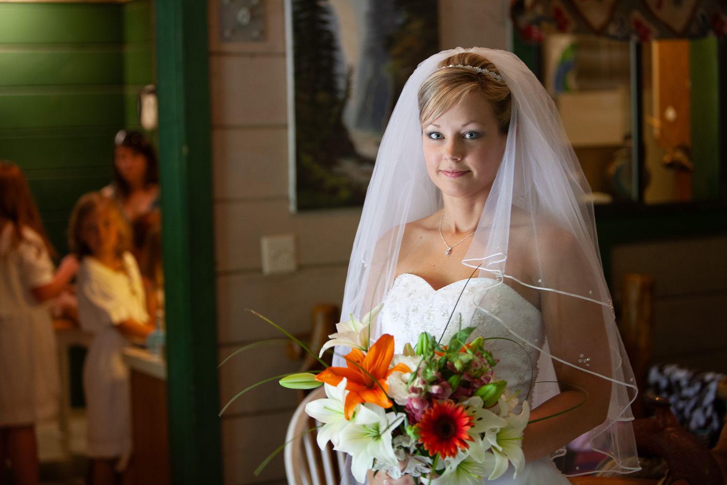 Elegant bride in a white gown and veil holding a colorful bouquet inside a warmly lit venue.