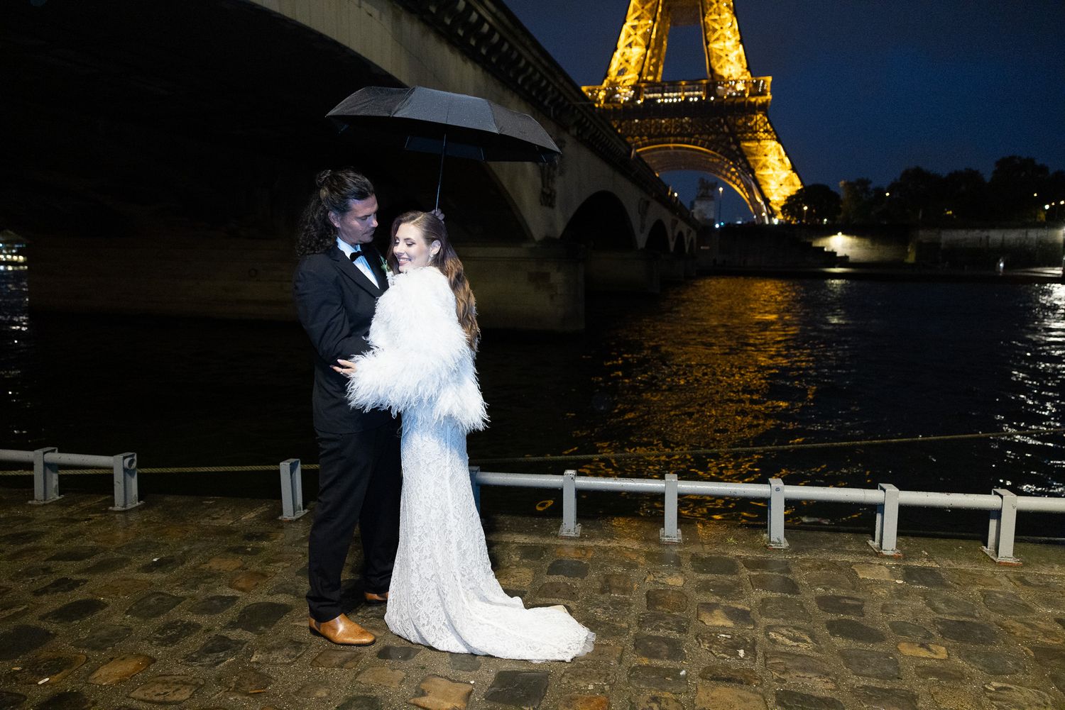 Newlyweds sharing an intimate moment under umbrella by the illuminated Eiffel Tower at night.