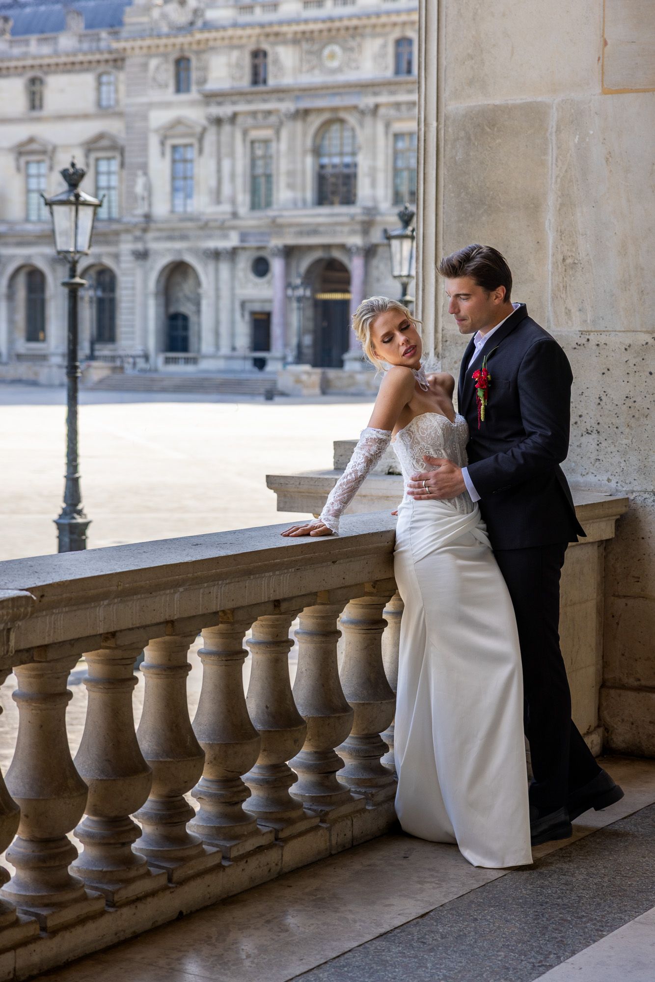 An elegant bride and groom pose together on a stone balcony at the Louvre in Paris.