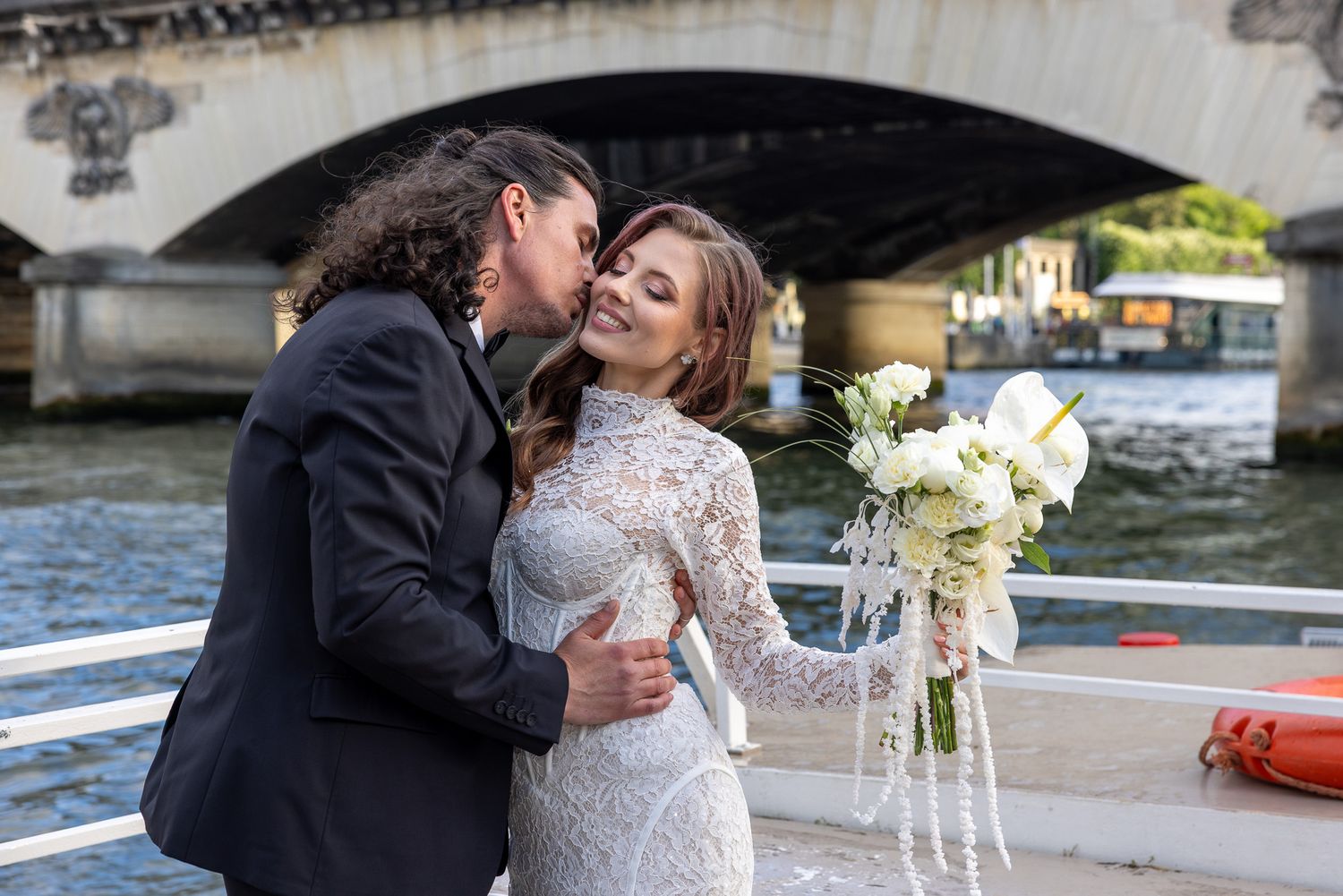 Newlyweds sharing a romantic kiss on a boat with a bridge in Paris background.