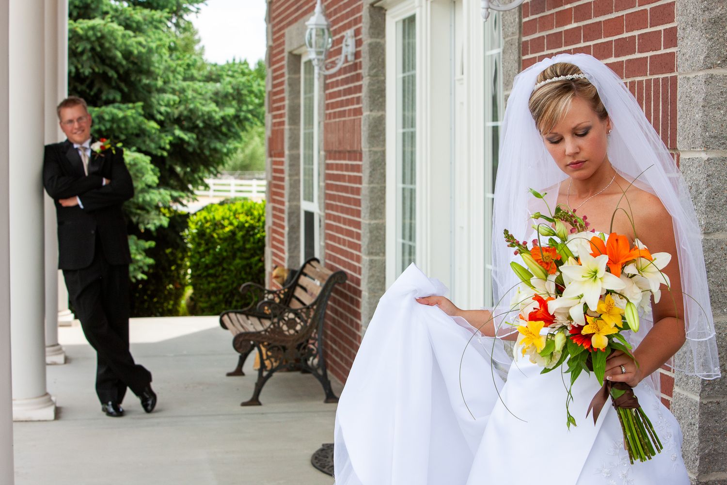 Bride holding colorful bouquet on church porch while groom approaches in background.