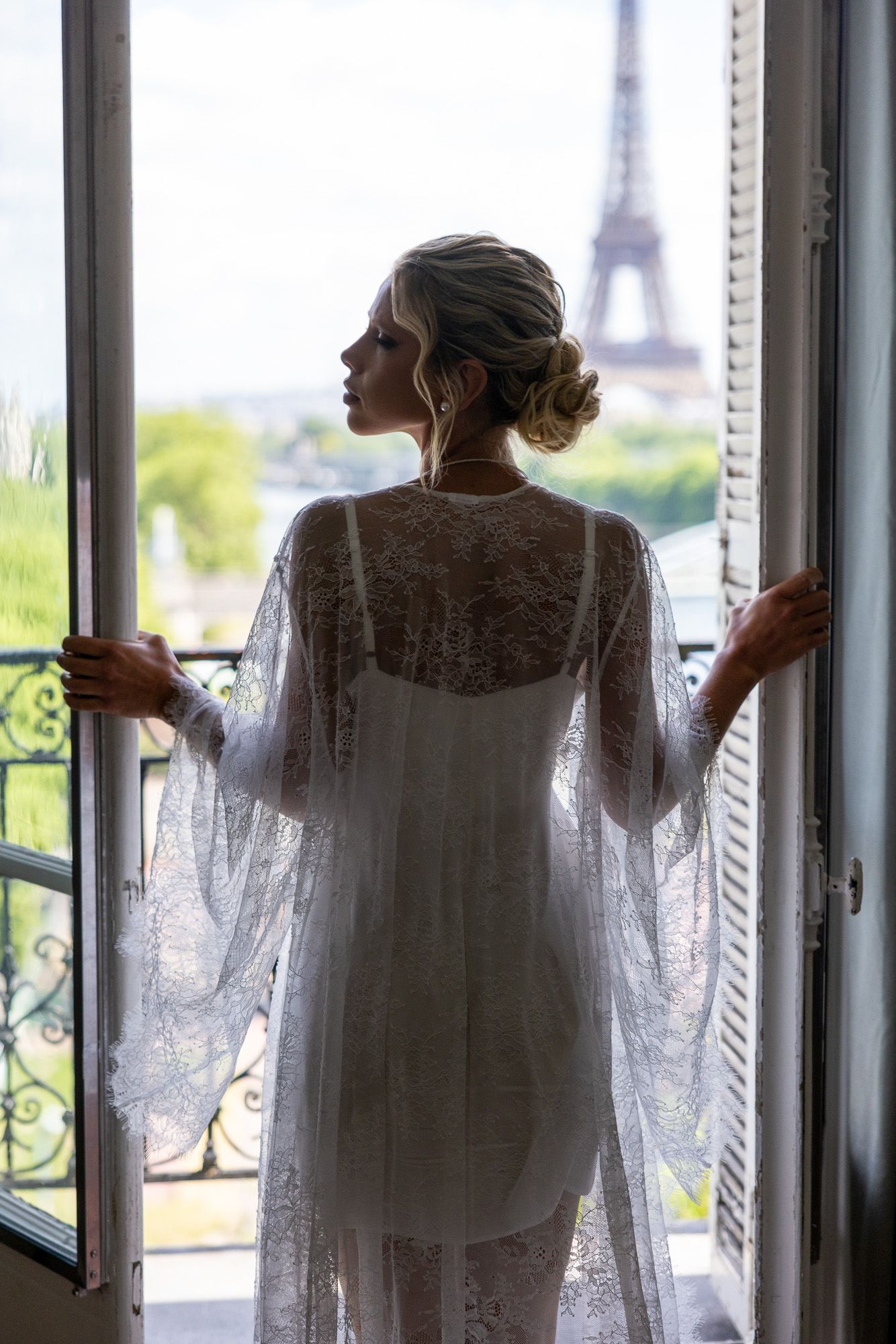 Bride in white lace robe opening balcony doors to reveal the iconic Eiffel Tower.
