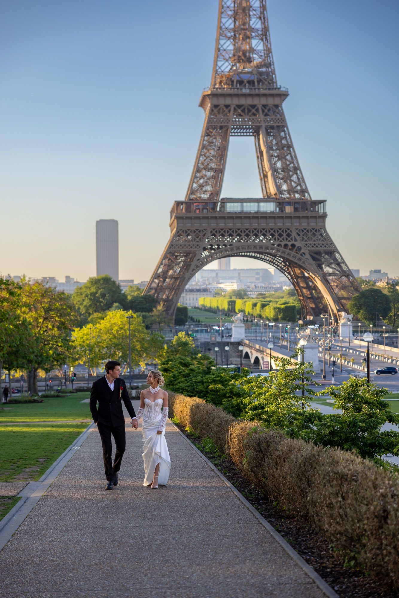 Bride and groom walk together on a sunlit path with the Eiffel Tower in background.
