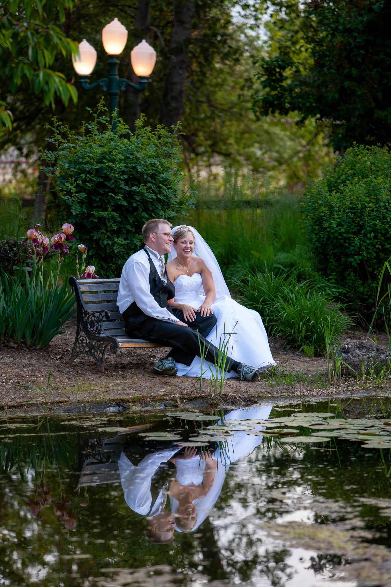 Romantic bride and groom cuddling on a garden bench by a lily pond at dusk with glowing street lamps.