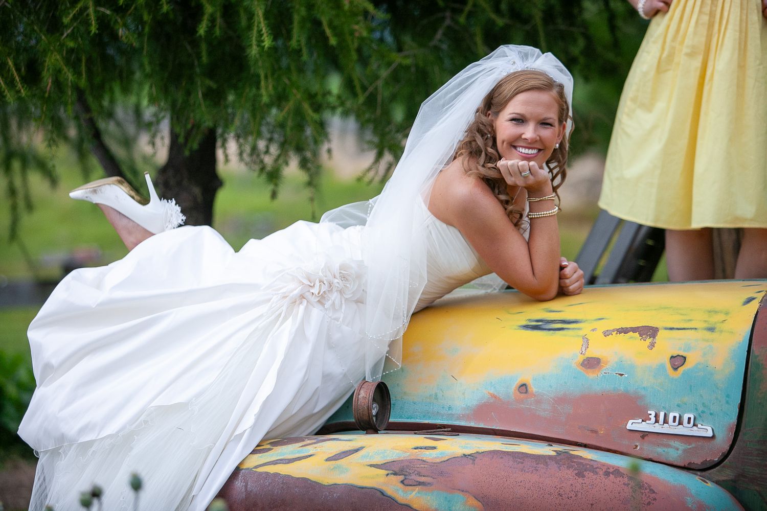 Radiant bride in white wedding gown leans on a colorful vintage truck hood smiling outdoors.