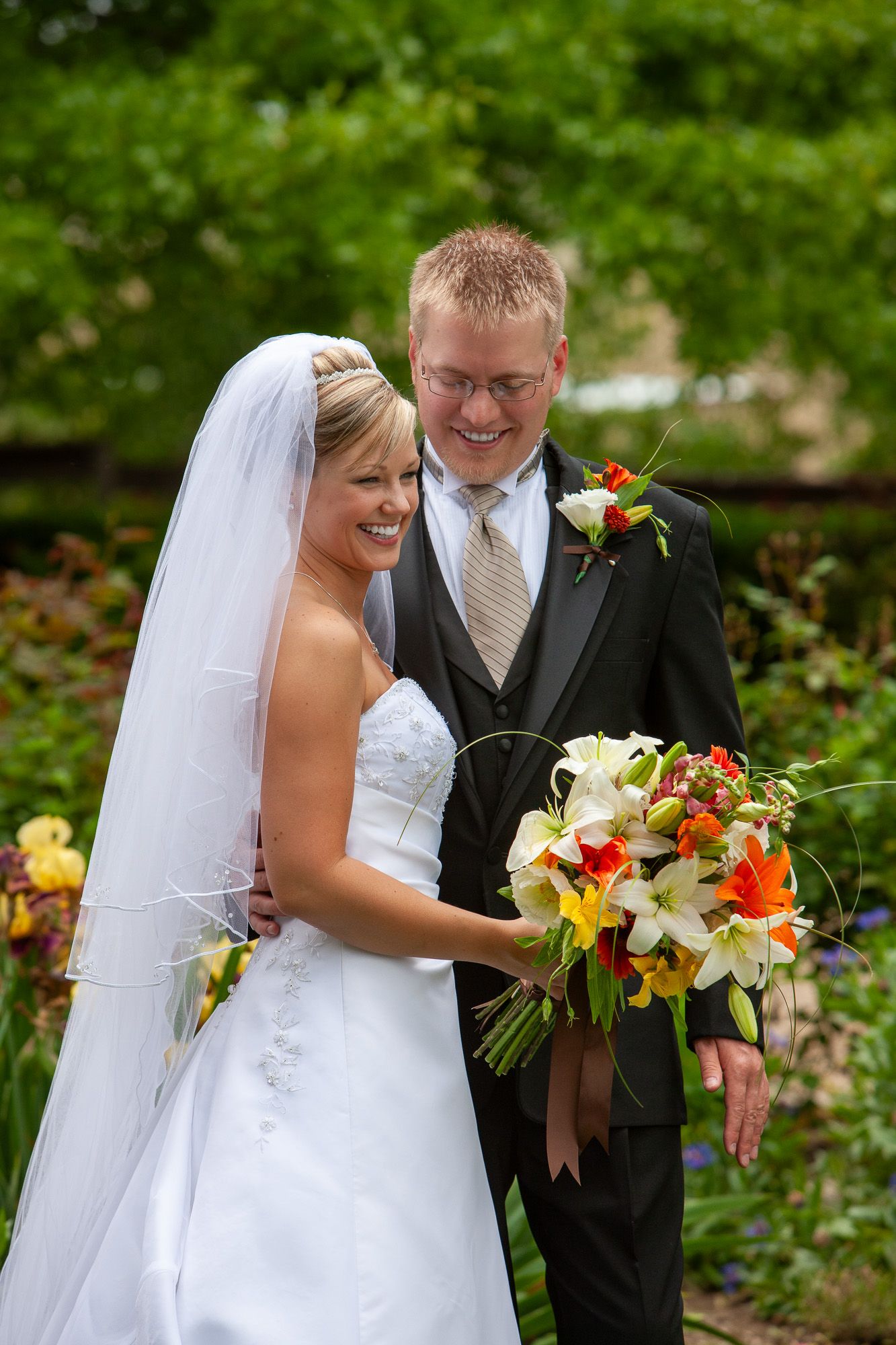 Smiling bride and groom posing together in garden holding colorful wedding bouquet.