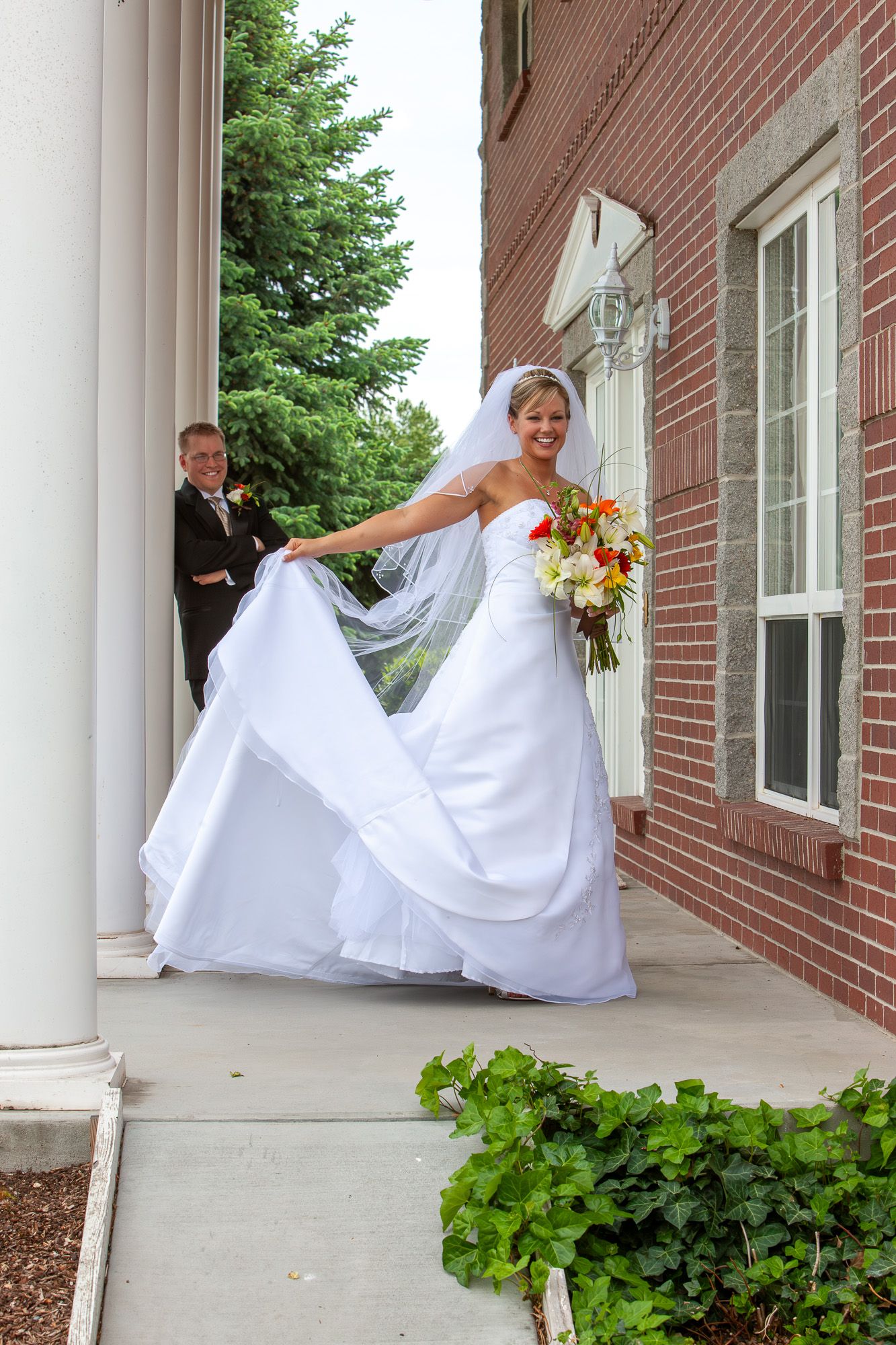 Smiling bride in white gown holding dress train outside brick church with groom.