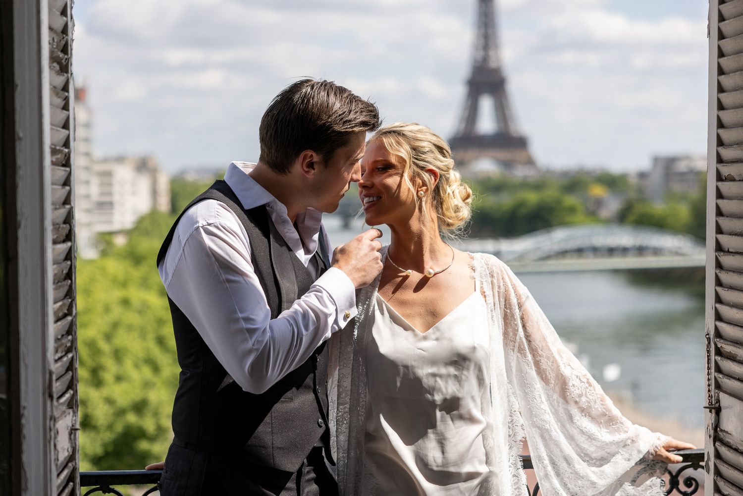 Groom kisses smiling bride on balcony with Eiffel Tower backdrop in Paris.