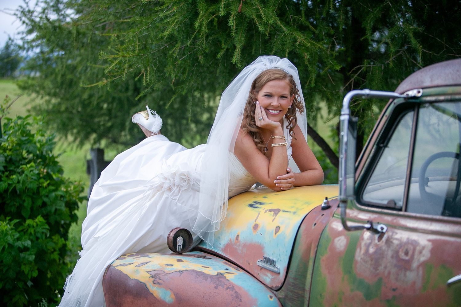 Smiling bride in white gown and veil poses on a rustic vintage truck outdoors near green trees.