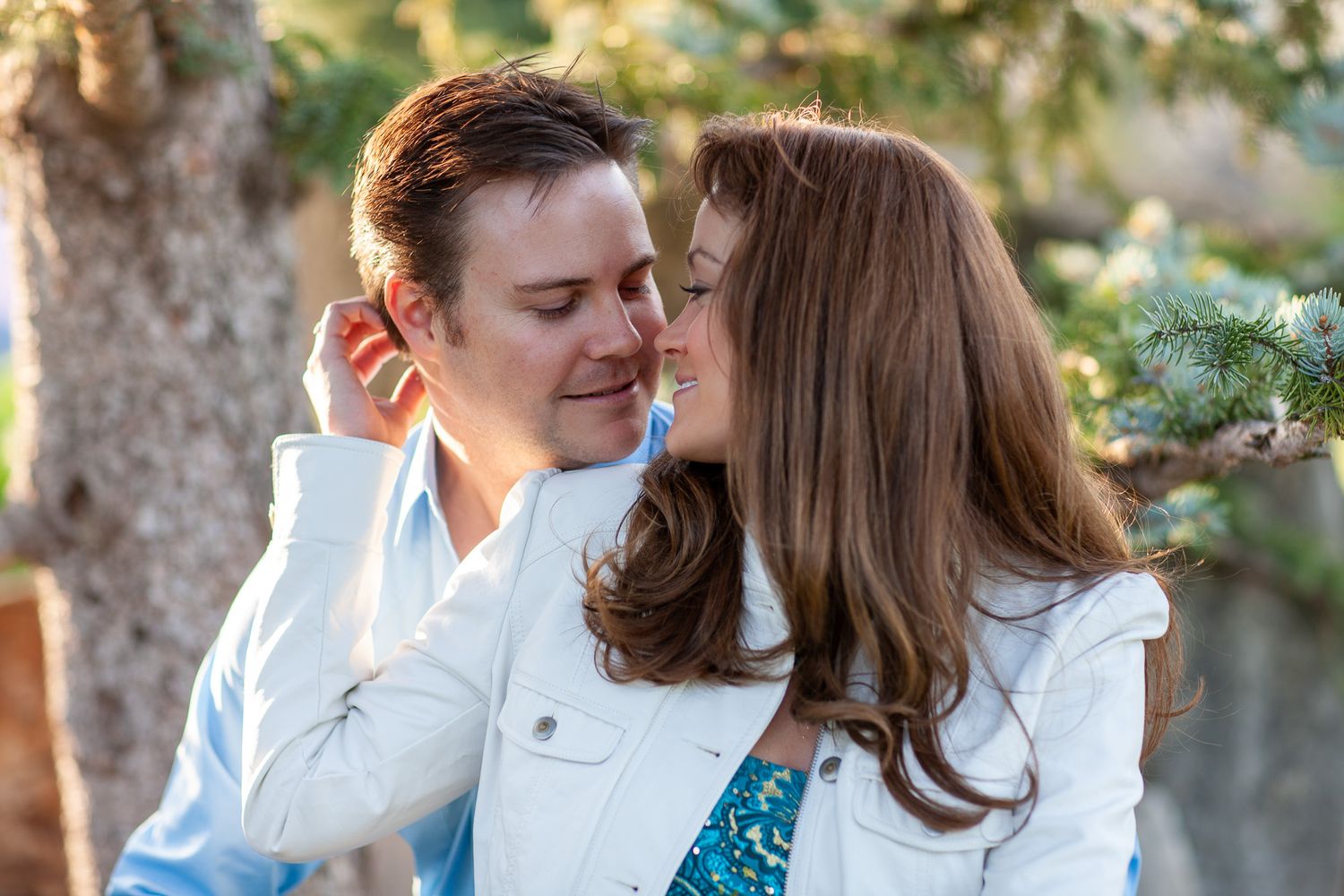 A woman gently kisses a mans cheek as they share a romantic moment outdoors near pine trees.