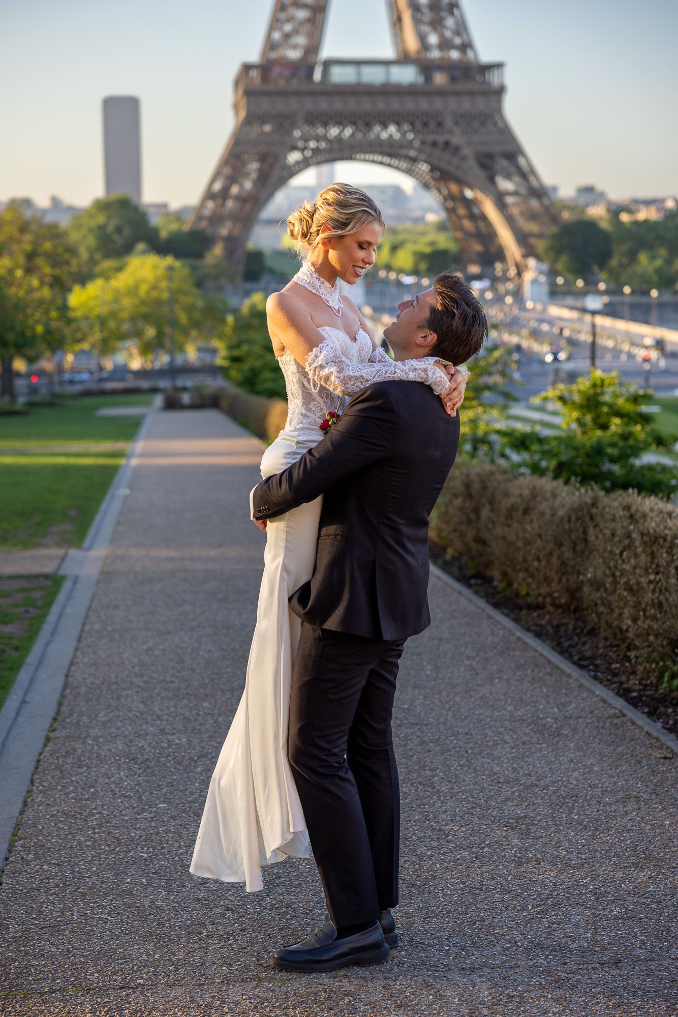Groom lifting bride in white lace gown near Eiffel Tower Paris at golden hour.
