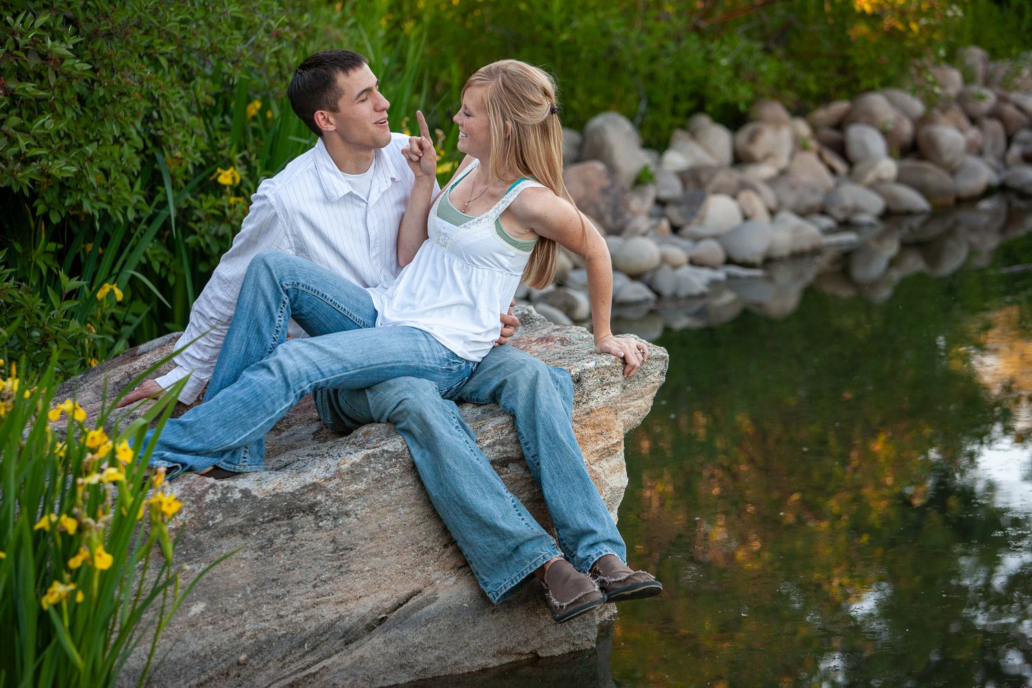 A young couple sitting on a large rock beside a calm pond surrounded by yellow irises and greenery.