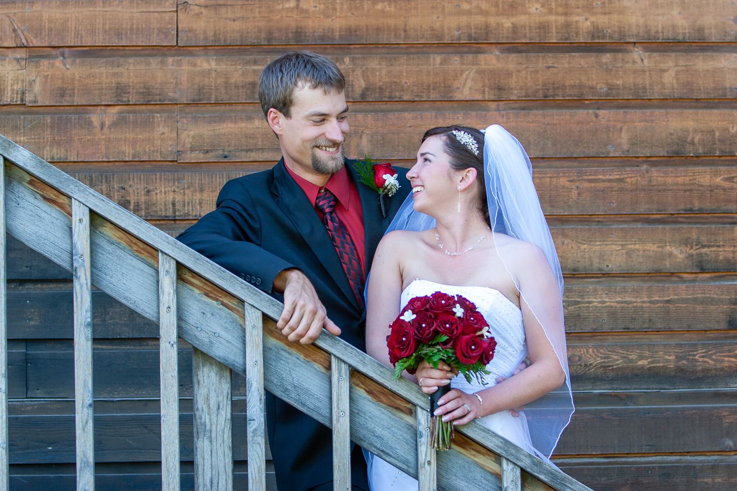 Bride and groom smiling at each other on wooden stairs, bride holding red roses bouquet on their wedding day.