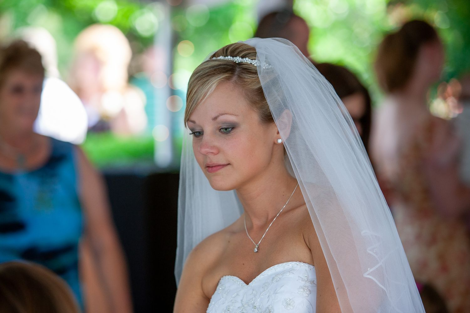 Beautiful bride in white gown and veil smiles softly during outdoor wedding ceremony.