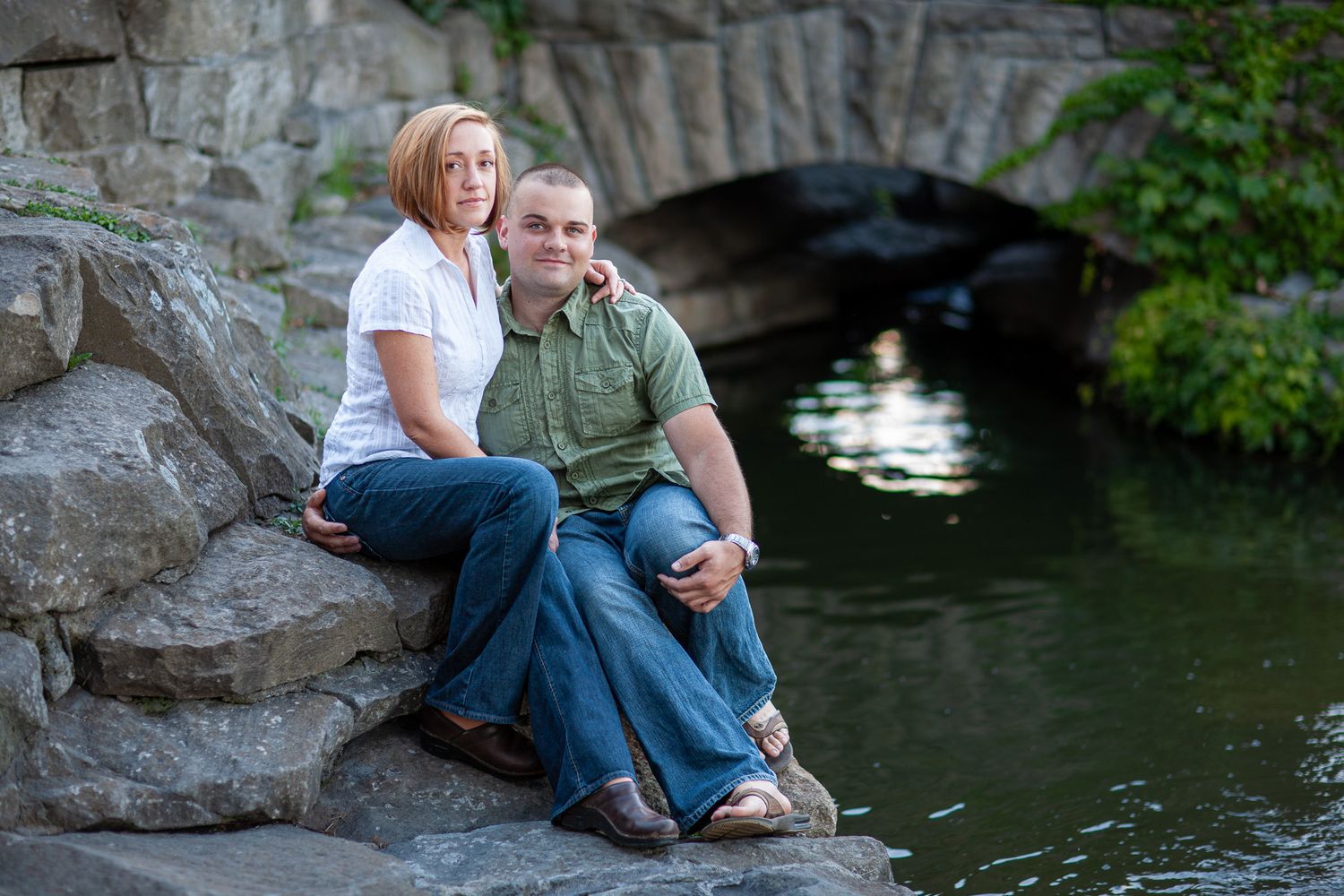 A couple sits together on rocks beside a tranquil pond with a stone bridge in the background during an engagement photo session.