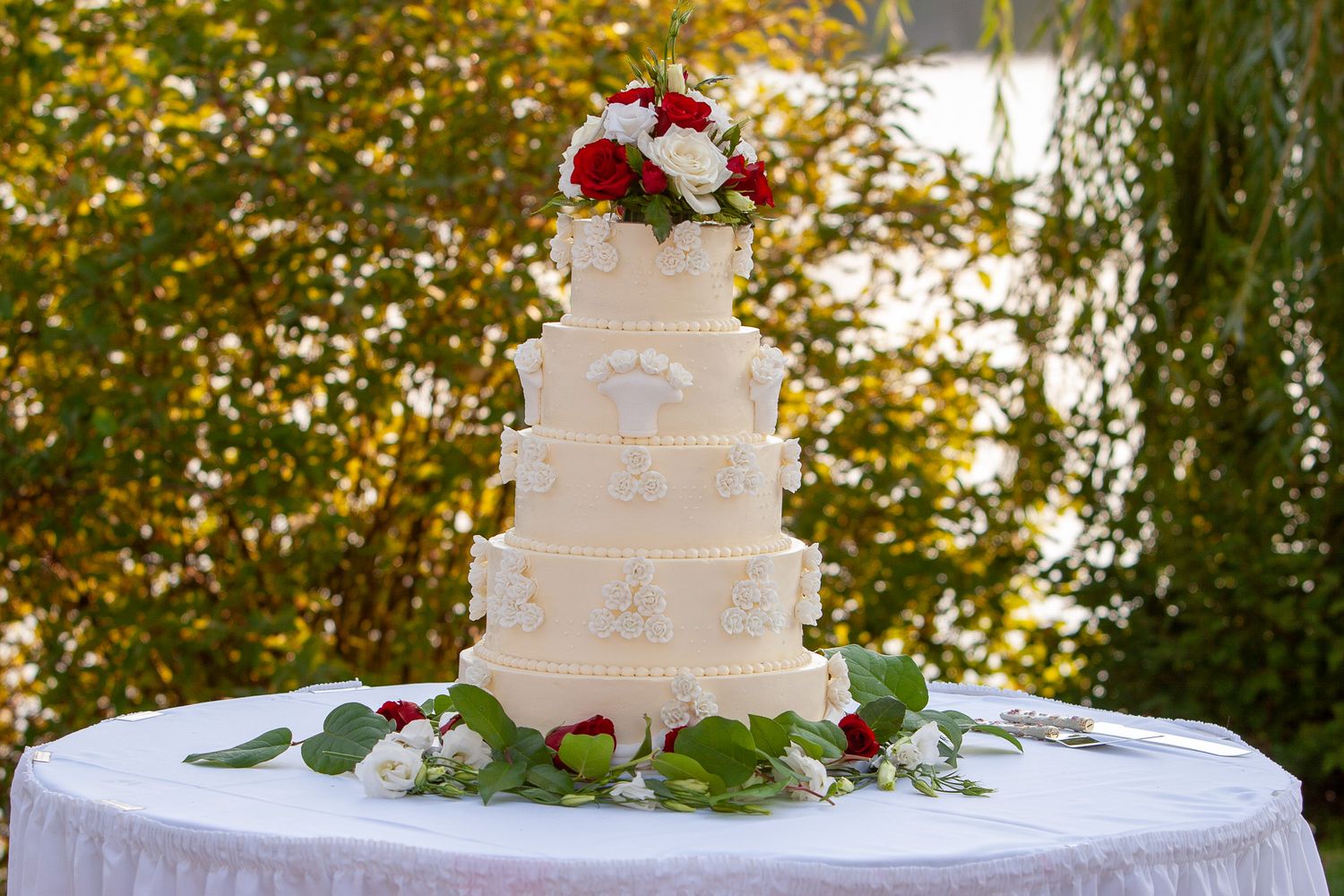 Elegant four-tier white wedding cake decorated with red and white roses outdoors.
