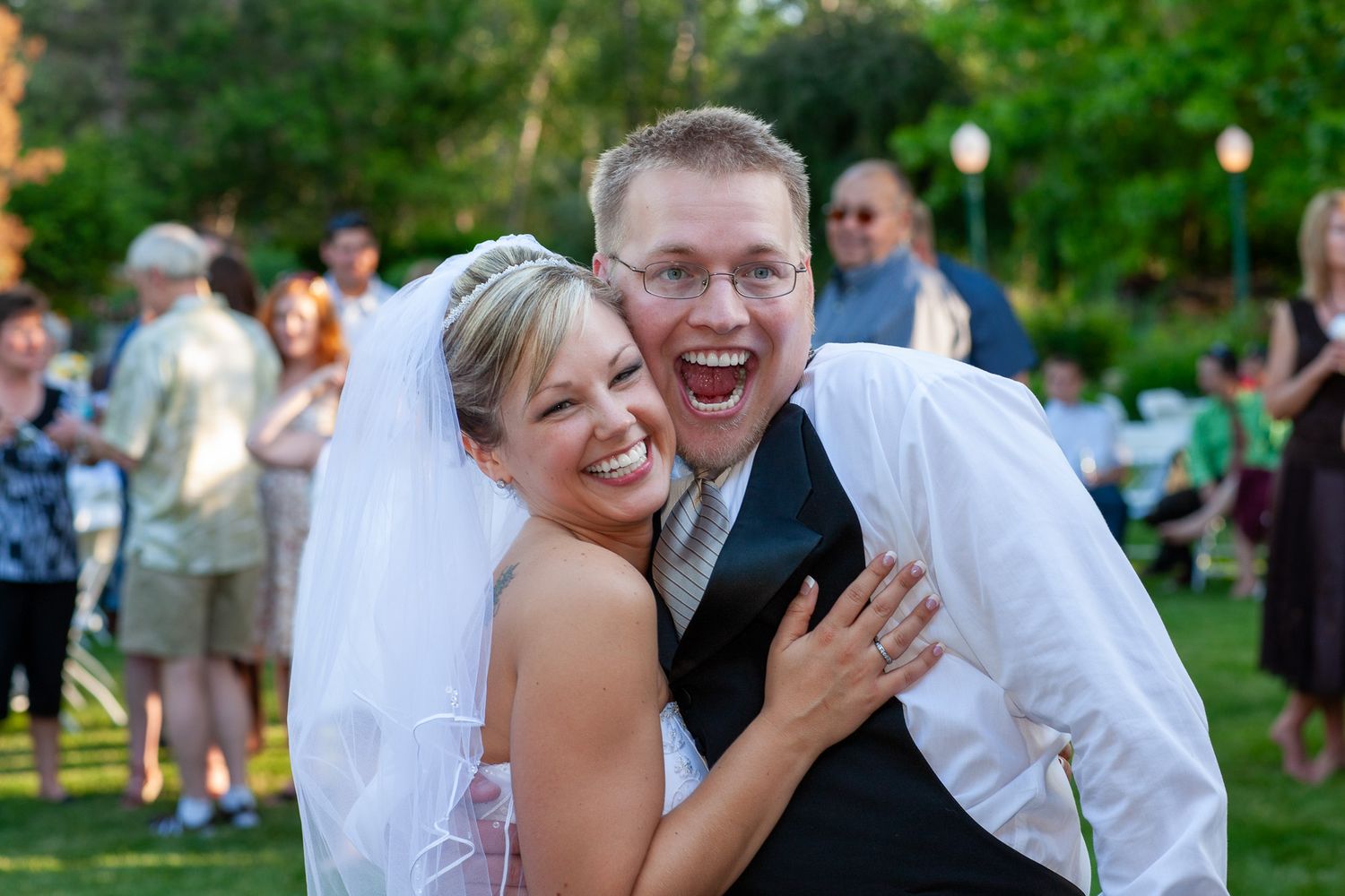 Joyful bride and groom laughing and embracing outdoors at their wedding reception in a lush green garden.
