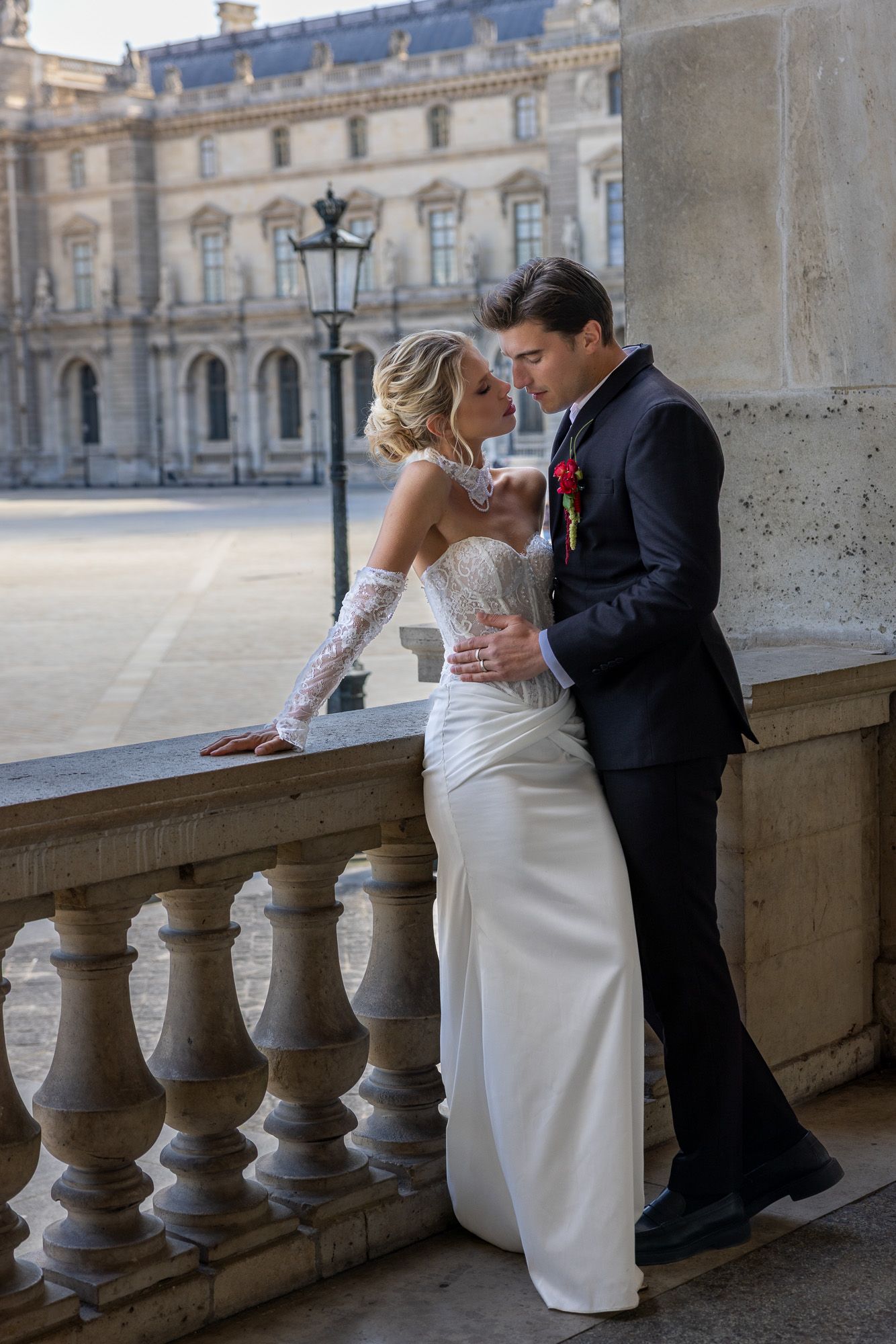 A bride in a white gown and groom in a dark suit pose on a stone balcony in Paris.