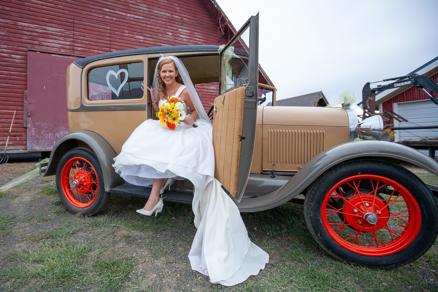 Smiling bride in white dress with yellow bouquet sits in a vintage tan car door near a red barn.