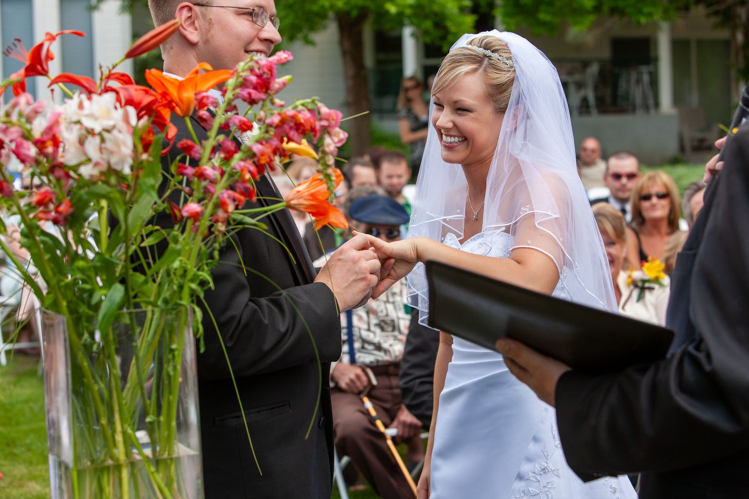 Bride laughing joyfully during outdoor wedding ceremony while exchanging rings with groom.