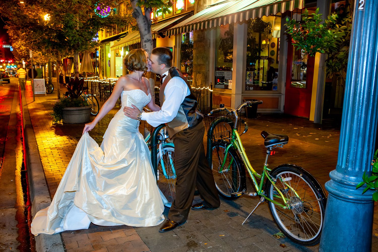Bride and groom sharing a romantic kiss on a charming city sidewalk at night near bicycles.