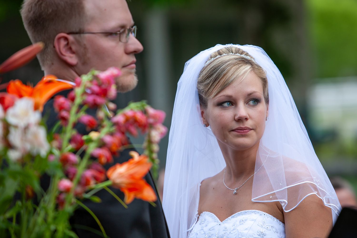Bride in white gown and veil gazes thoughtfully during outdoor wedding ceremony with colorful flowers nearby.