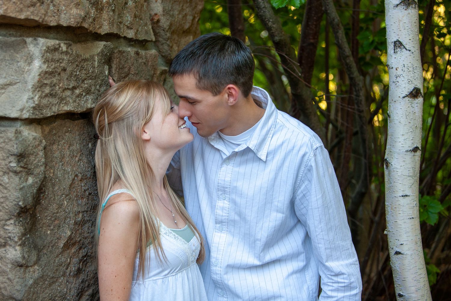 A young blonde woman and a man share a sweet nose-to-nose moment against a stone wall outdoors.