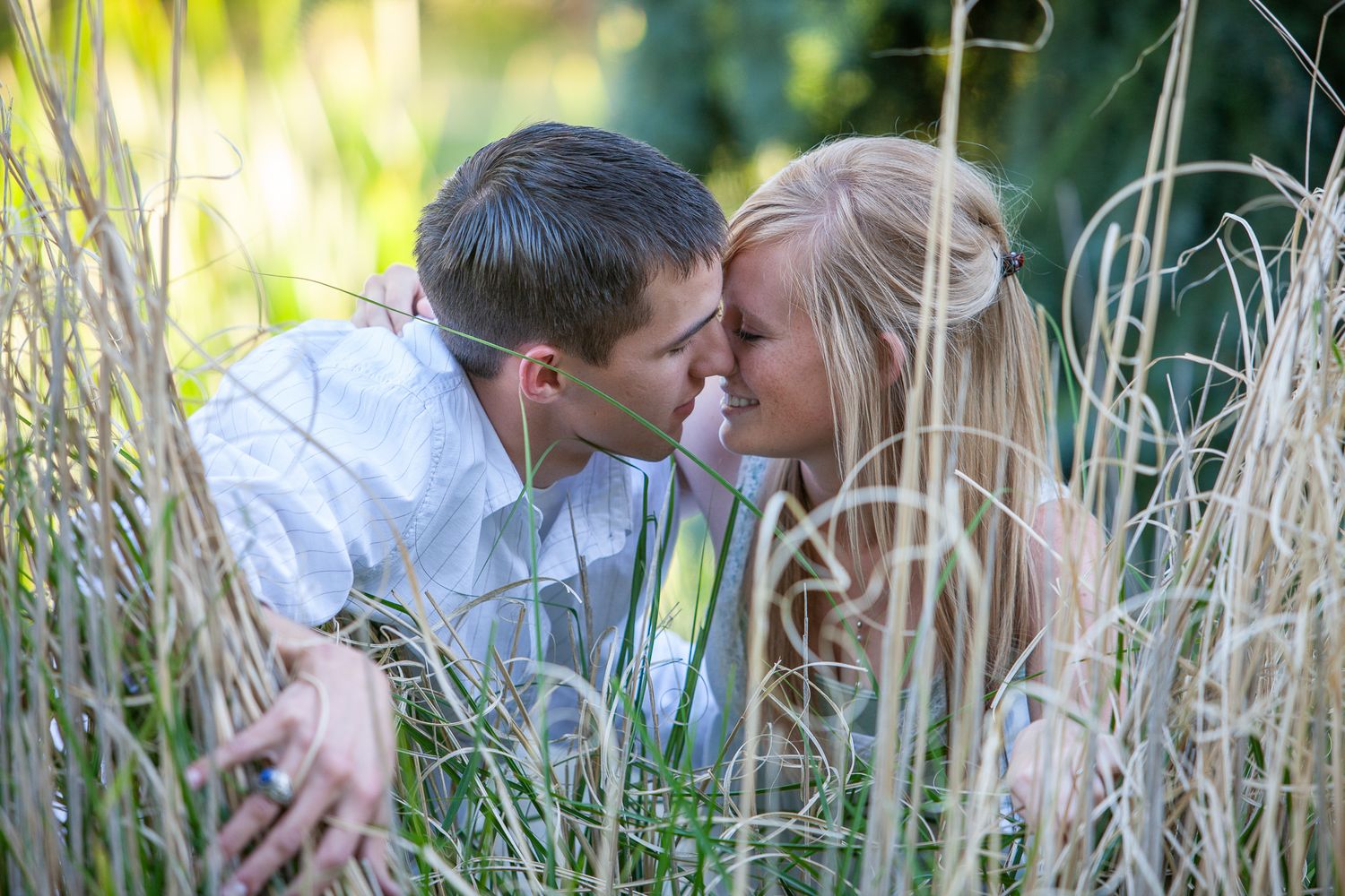 A romantic couple sharing an intimate moment touching foreheads while nestled among tall green grass outdoors.